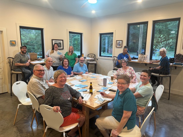 thirteen people sitting around a square table writing postcards